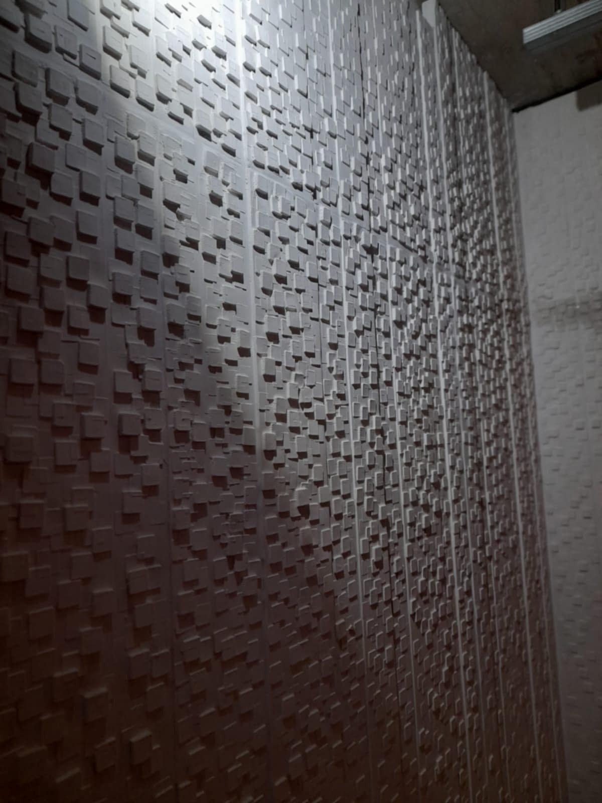 Ściana pokryta trójwymiarowymi, małymi kwadratami, tworzącymi geometryczną fakturę. Widoczne fragmenty sufitu w tle.