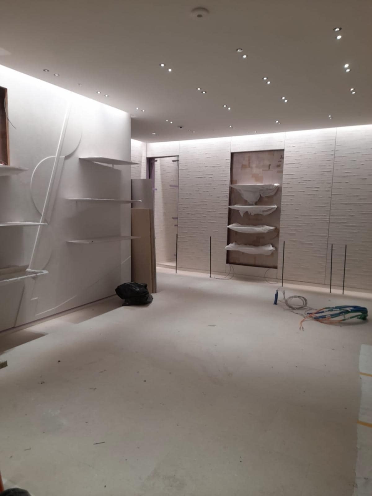 Wnętrze sklepu w trakcie wykańczania, widoczne regały i ściany z dekoracyjną fakturą, przewody elektryczne na podłodze, oświetlenie punktowe w suficie.