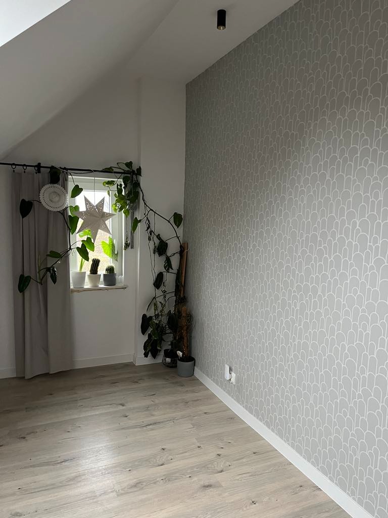 Pokój na poddaszu po remoncie: szare tapety w subtelny wzór, jasna podłoga imitująca drewno, okno z dekoracjami roślinnymi i gwiazdą, minimalistyczne oświetlenie.
