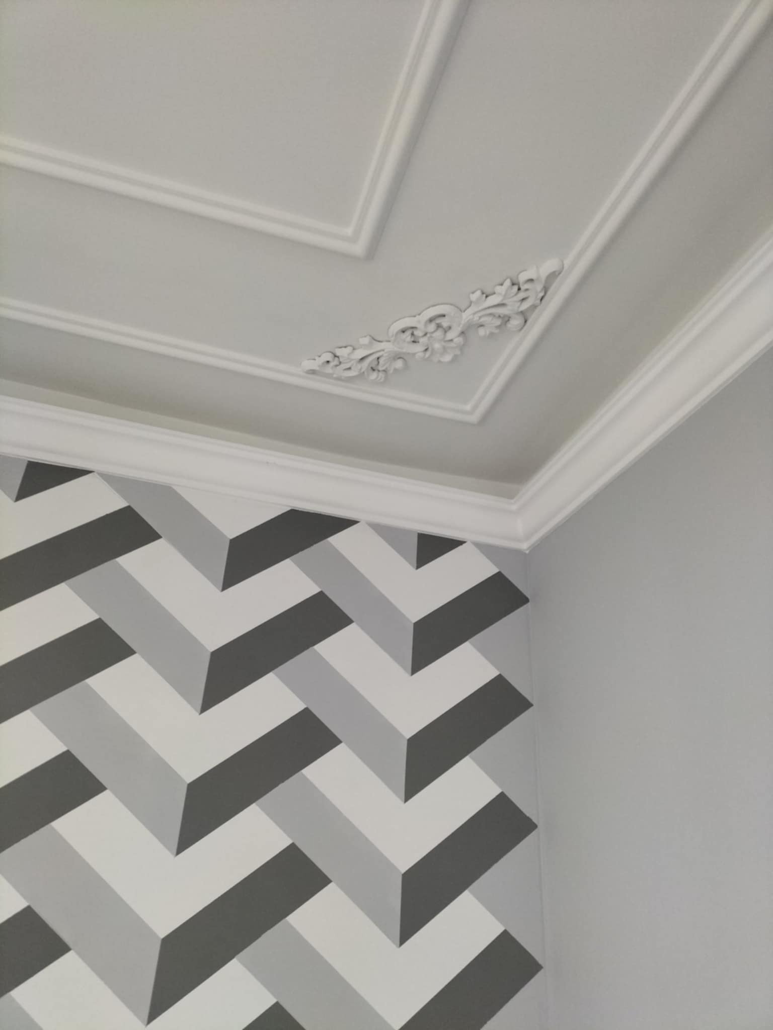 Wykończony róg pokoju z dekoracyjnym sztukateryjnym sufitem, ozdobnym elementem narożnym i ścianą z geometrycznym wzorem w odcieniach szarości.