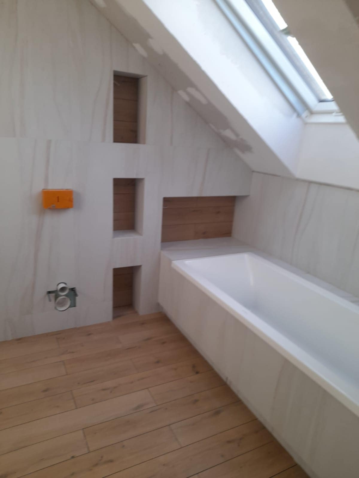 Łazienka na poddaszu z drewnopodobnymi płytkami podłogowymi, zabudowaną wanną, płytkami ściennymi imitującymi kamień i wnękami wykończonymi drewnem.