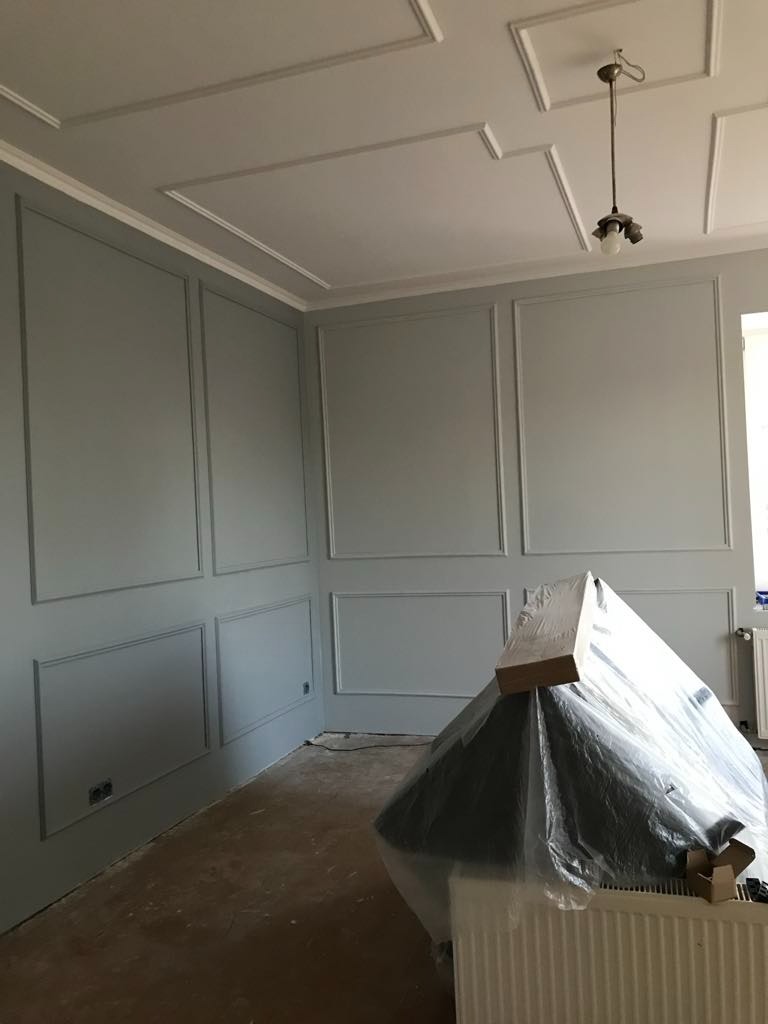 Pokój po częściowym odświeżeniu, ściany w odcieniach szarości z dekoracyjnymi listwami, sufit z geometrycznymi zdobieniami, zakryty mebel w folii ochronnej, widoczny grzejnik.