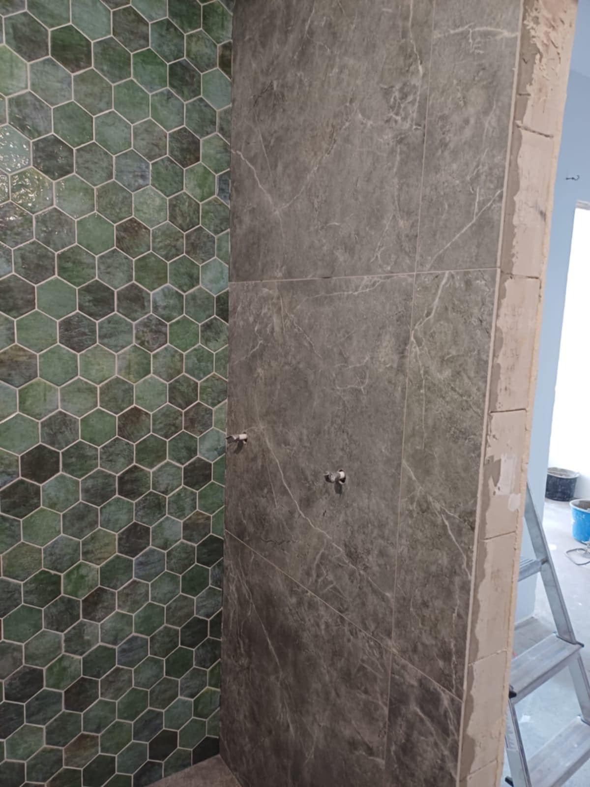 Wykończona ściana prysznica w Oławie z heksagonalnymi, zielonymi płytkami po lewej i szarymi płytkami imitującymi marmur po prawej, z widocznymi przyłączami do baterii i fragmentem drabiny malarskiej.