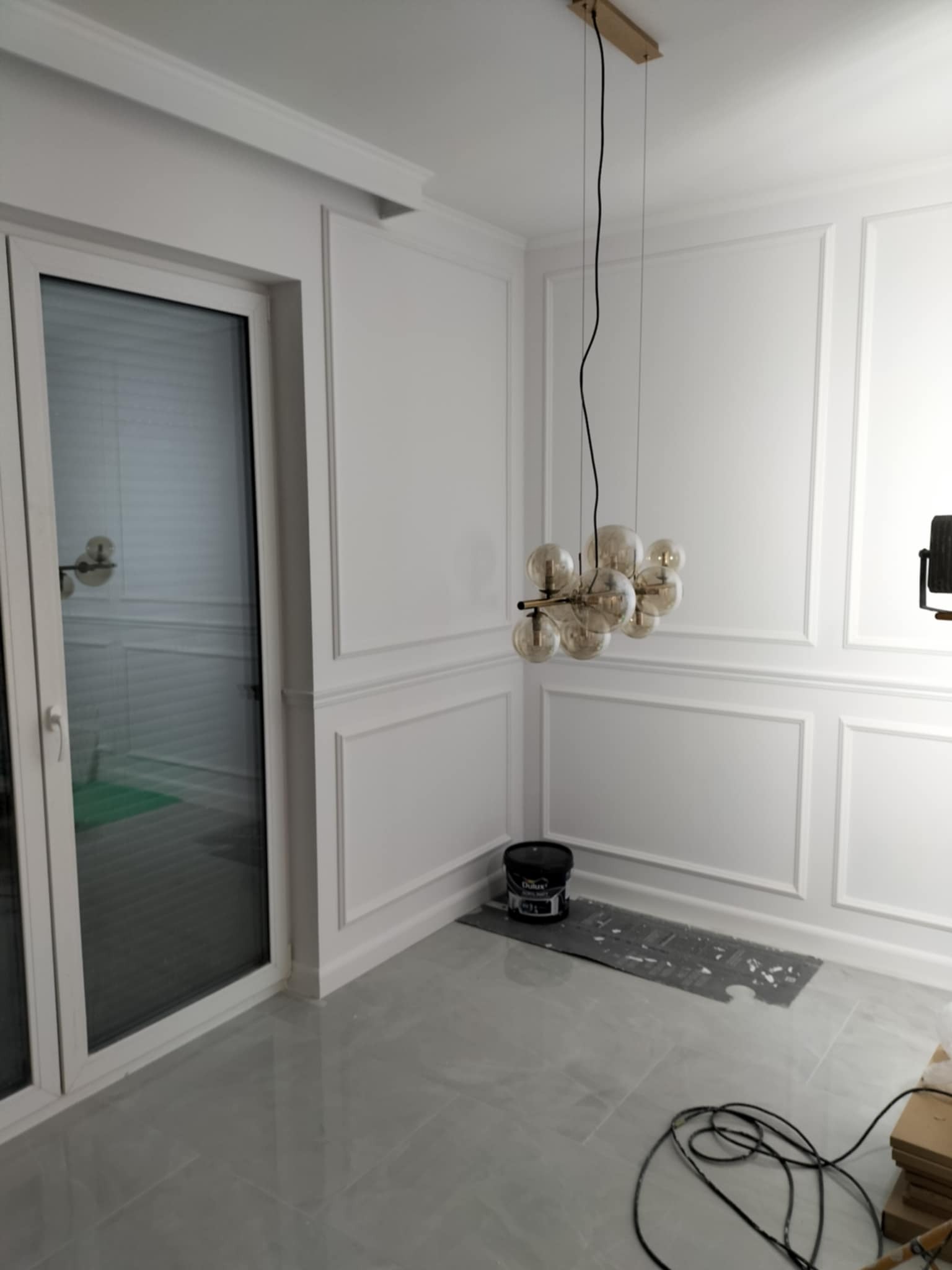 Wykończone wnętrze w stylu klasycznym z białymi ścianami zdobionymi sztukaterią, szarą podłogą z płytek, nowoczesną lampą wiszącą z kulistych kloszy i widocznym wiadrem z farbą Dulux oraz kablami...