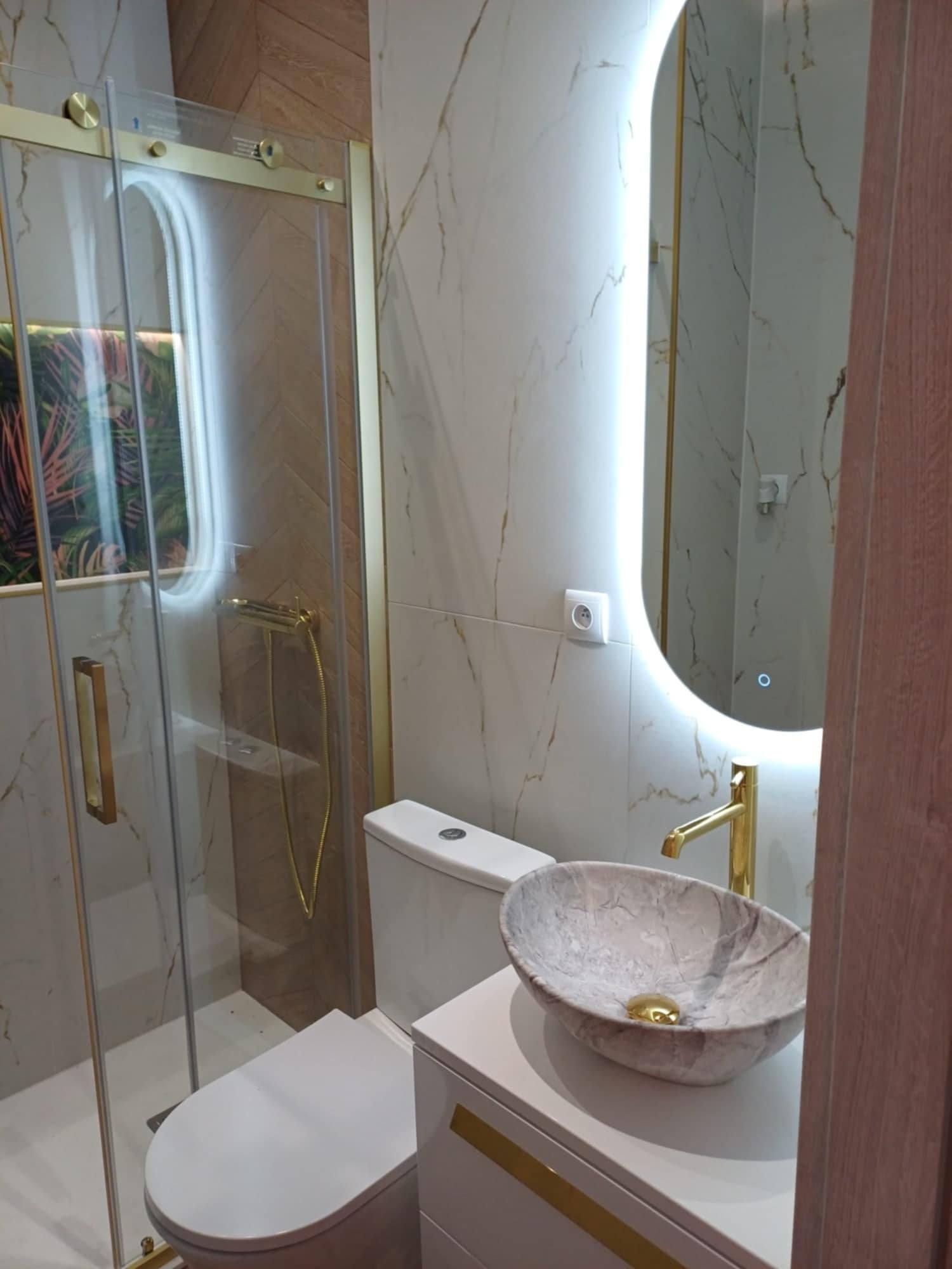 Nowoczesna łazienka z marmurową umywalką nablatową, złotą armaturą, podświetlanym lustrem i kabiną prysznicową ze złotymi detalami.