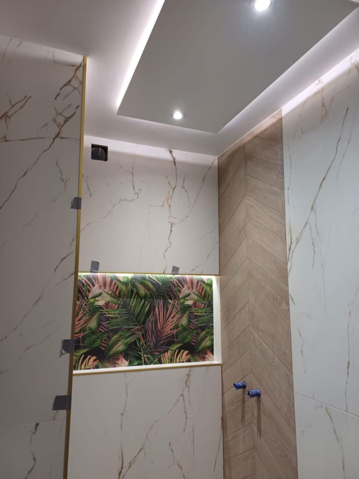 Wnętrze łazienki w trakcie wykańczania, z białymi marmurkowymi płytkami, drewnopodobną ścianą ułożoną w jodełkę, wnęką z motywem roślinnym i podwieszanym sufitem z oświetleniem LED.