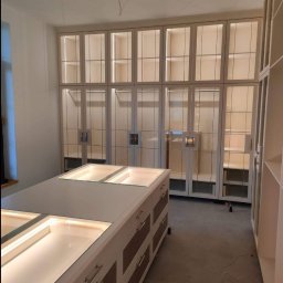 Styl mebel - Jasna garderoba z przeszklonymi szafami i wyspą z szufladami, podświetlona ledami, widoczne kable na suficie, w trakcie montażu.