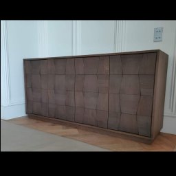 Styl mebel - Długa, niska szafka z frontami o geometrycznym wzorze, wykonana z drewna w odcieniach szarości, ustawiona na parkiecie w jasnym wnętrzu z dekoracyjną sztukaterią.