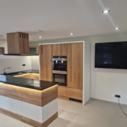 Nowoczesna kuchnia z wyspą, drewnianymi frontami, czarnym blatem i podświetleniem LED, zabudowana szafa z piekarnikiem i ekspresem do kawy, minimalistyczny okap.