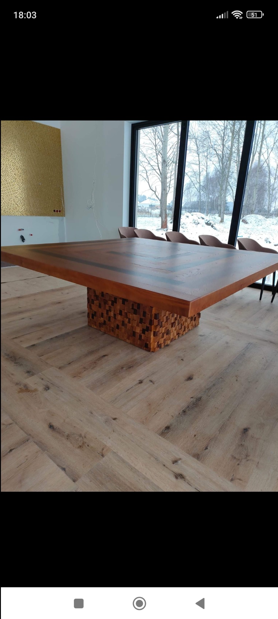 Duży, kwadratowy stół z drewnianym blatem i podstawą z mozaiki drewnianych kostek, ustawiony w jasnym pomieszczeniu z dużym oknem i drewnianą podłogą w jodełkę.