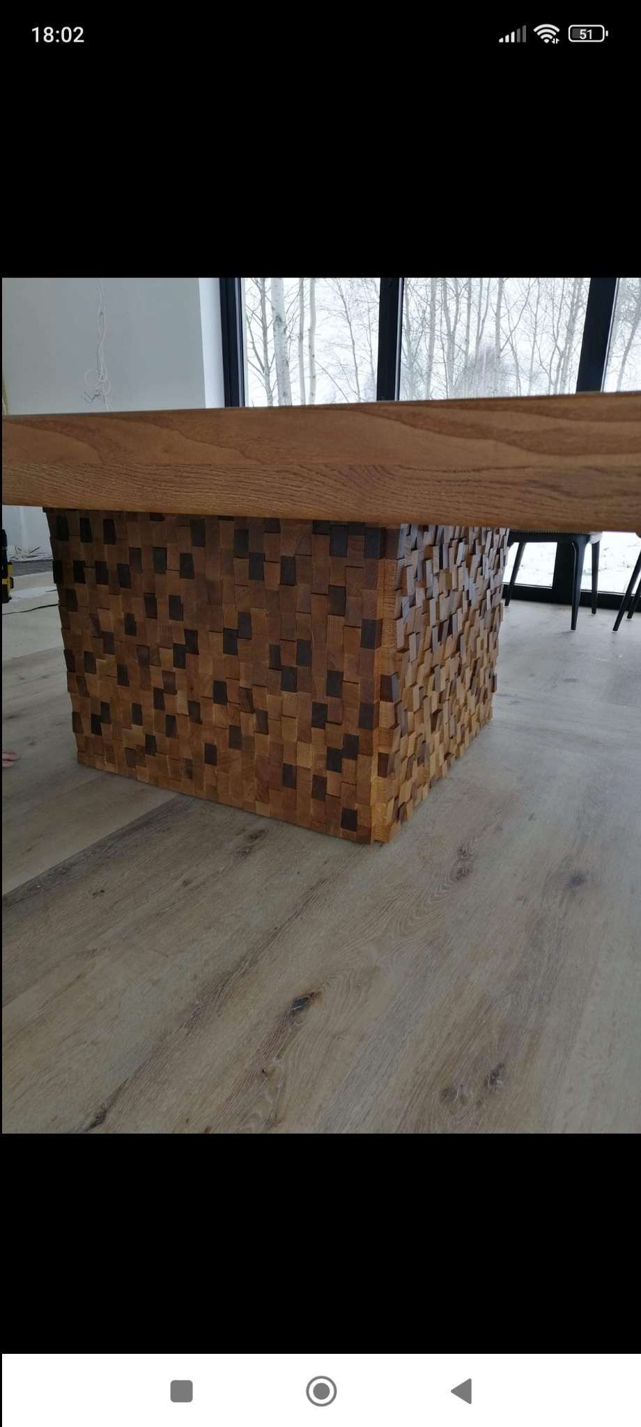 Stół z blatem z jasnego drewna i podstawą z mozaiki ciemniejszych drewnianych kostek, na tle okna z widokiem na drzewa.