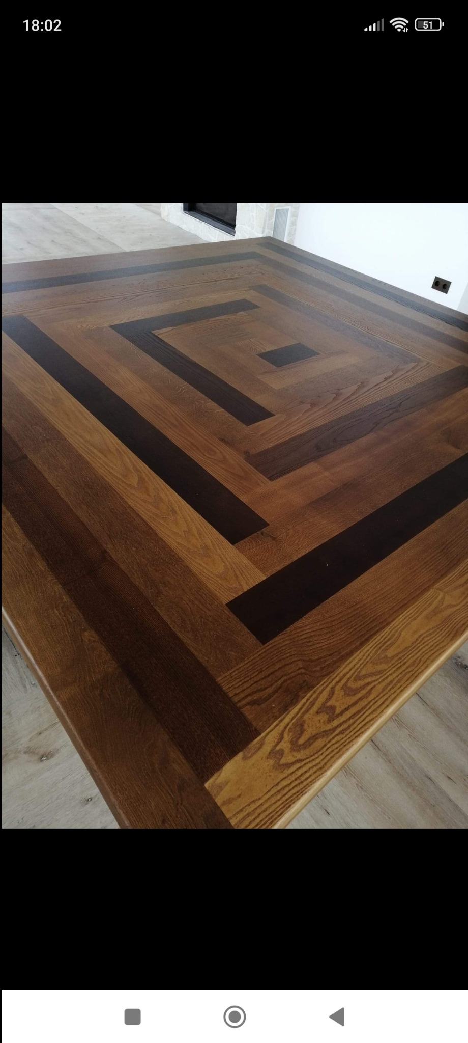 Parkiet ułożony w geometryczny wzór kwadratu z ciemnego i jasnego drewna, widoczny fragment ściany z gniazdkiem elektrycznym i kominkiem w tle.