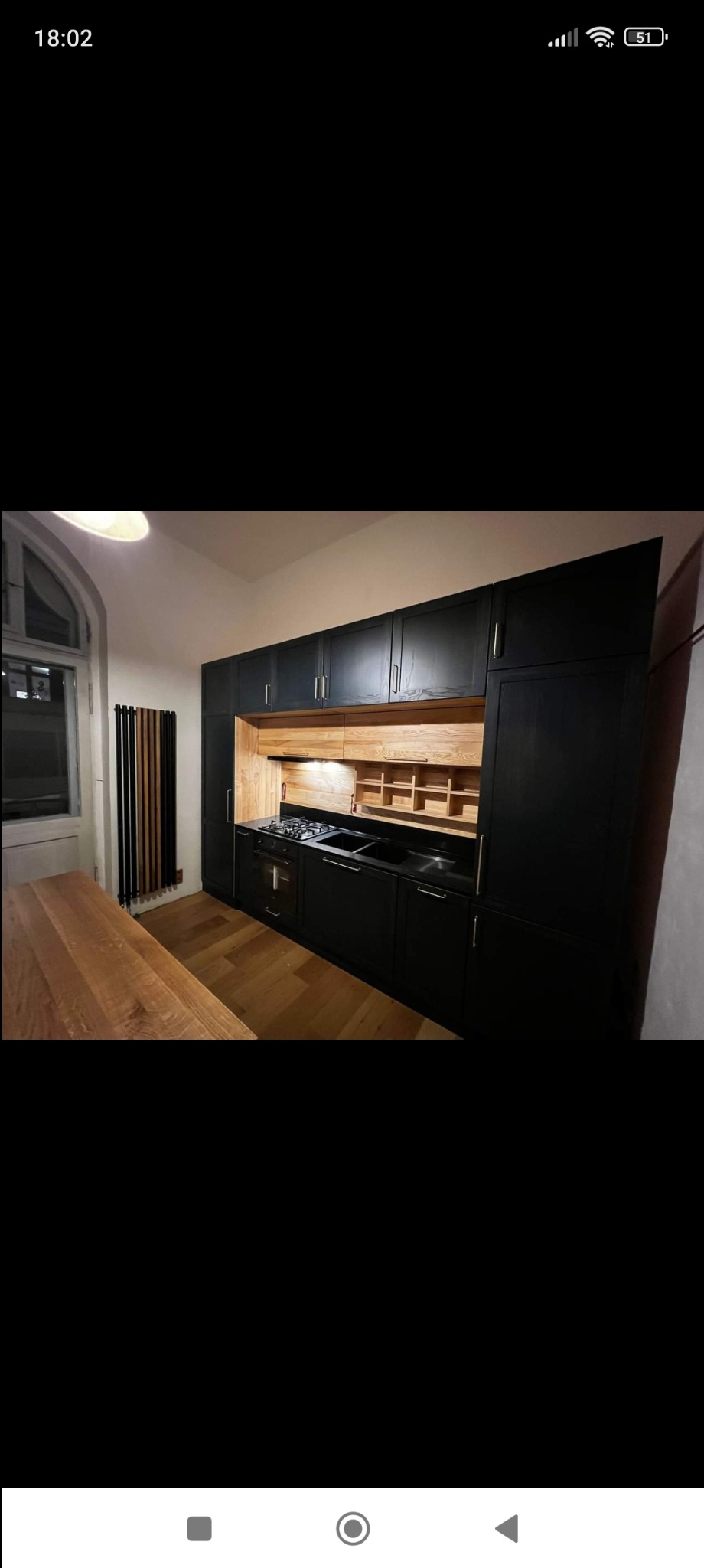 Ciemna zabudowa kuchenna z drewnianymi elementami, widoczny stół z tego samego drewna, grzejnik dekoracyjny w tle.