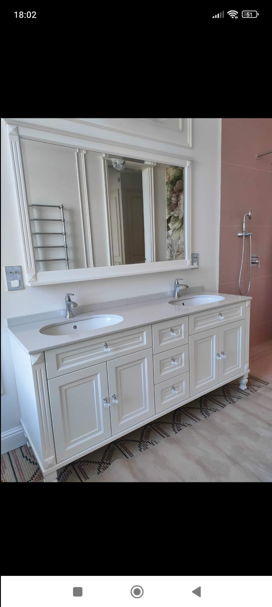 Elegancka, kremowa szafka łazienkowa z dwoma umywalkami nablatowymi i kryształowymi gałkami, zwieńczona dużym lustrem z dekoracyjną ramą. Widoczny fragment ściany z różowymi płytkami i prysznicem.