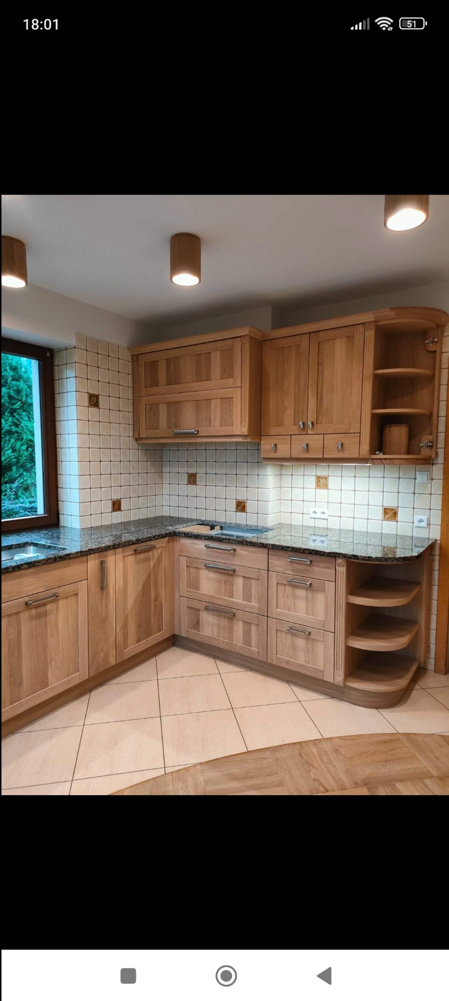 Jasna, drewniana zabudowa kuchenna narożna z blatem w kolorze ciemnego granitu, widoczne oświetlenie sufitowe i okno z zielenią na zewnątrz.