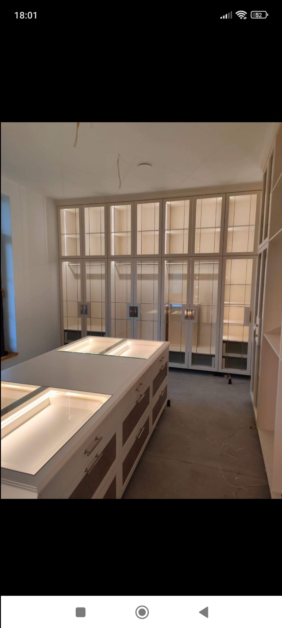 Jasna garderoba z przeszklonymi szafami i wyspą z szufladami, podświetlona ledami, widoczne kable na suficie, w trakcie montażu.