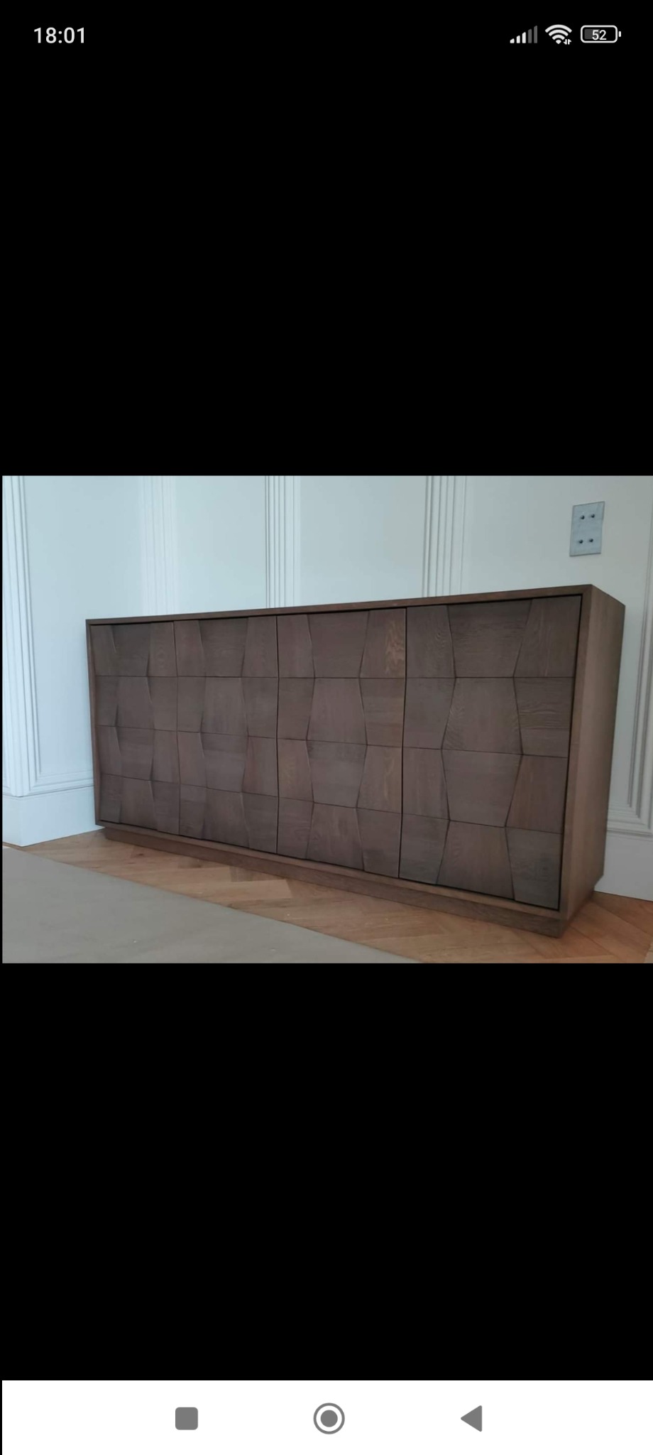 Długa, niska szafka z frontami o geometrycznym wzorze, wykonana z drewna w odcieniach szarości, ustawiona na parkiecie w jasnym wnętrzu z dekoracyjną sztukaterią.