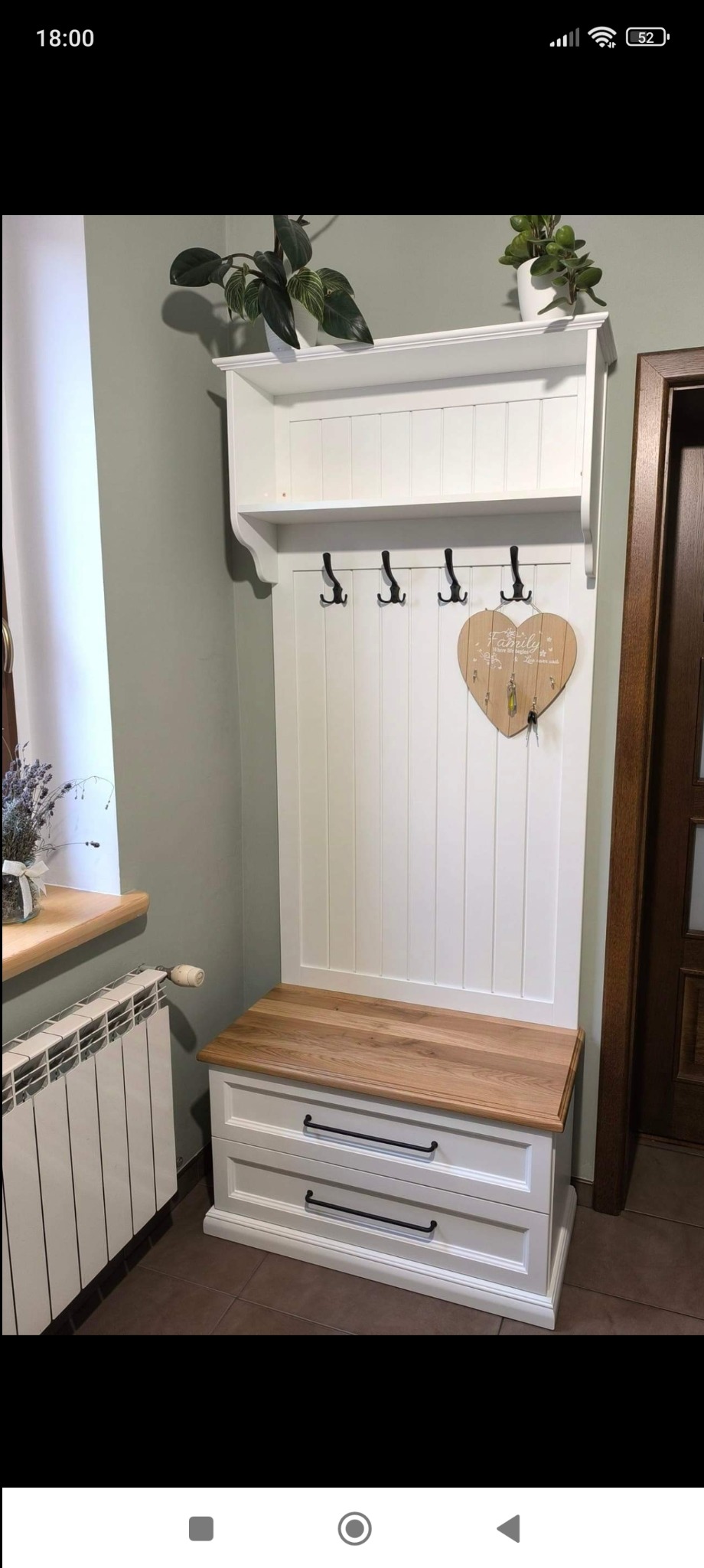 Biały mebel do przedpokoju z drewnianym siedziskiem, haczykami na ubrania i półką górną z roślinami, obok kaloryfer i drewniane drzwi.