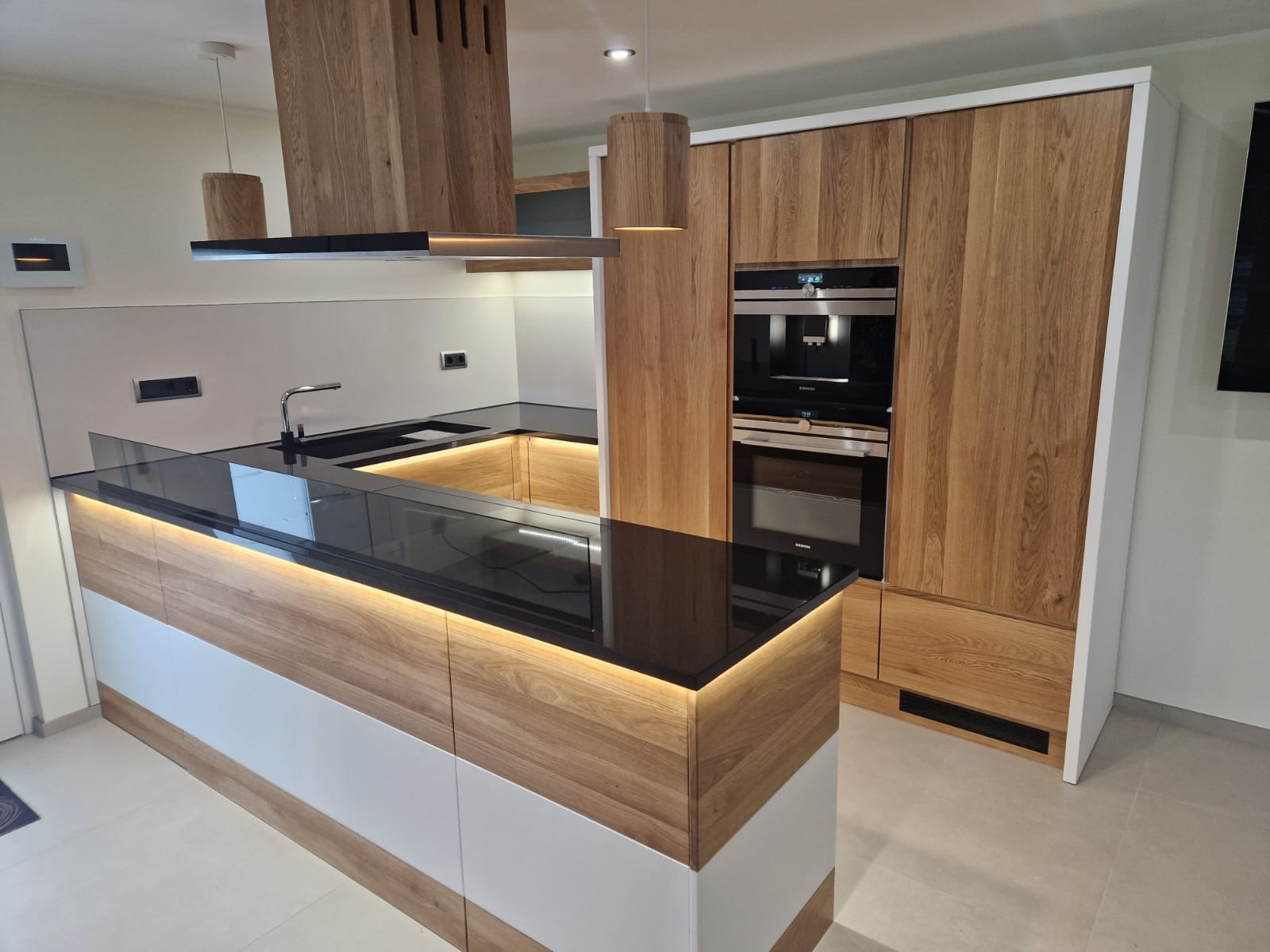 Nowoczesna kuchnia z drewnianymi frontami, czarnym blatem i oświetleniem LED pod szafkami, zabudowany ekspres do kawy i piekarnik w wysokiej szafce, drewniany okap nad wyspą.