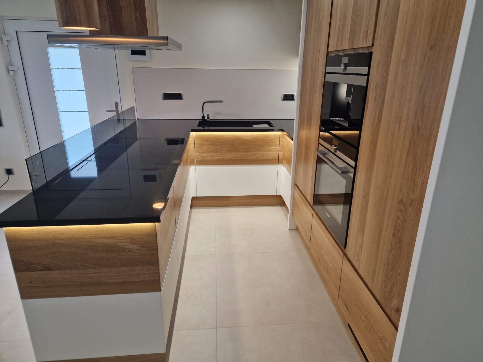 Nowoczesna kuchnia z drewnianymi frontami szafek, czarnym blatem i podświetleniem LED, z widocznym piekarnikiem w zabudowie.