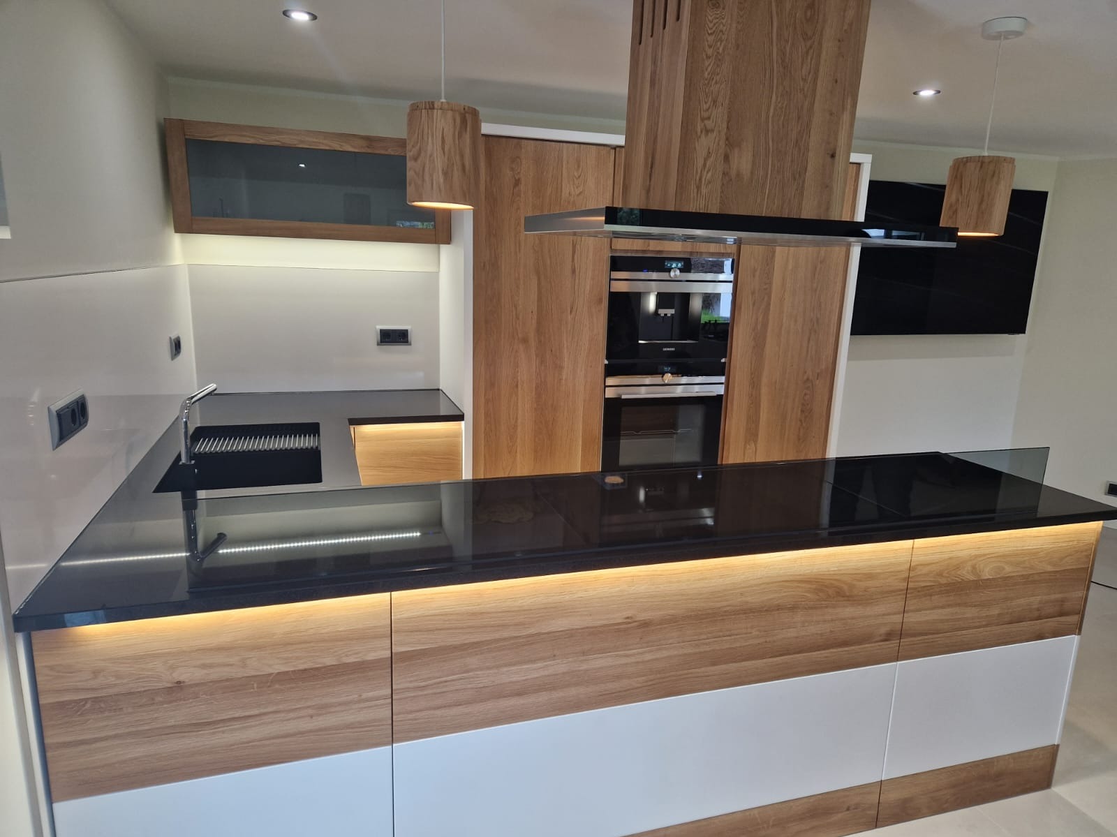 Nowoczesna kuchnia z wyspą, drewnianymi frontami szafek dolnych z podświetleniem LED, czarnym blatem i wiszącymi lampami z drewnianymi abażurami