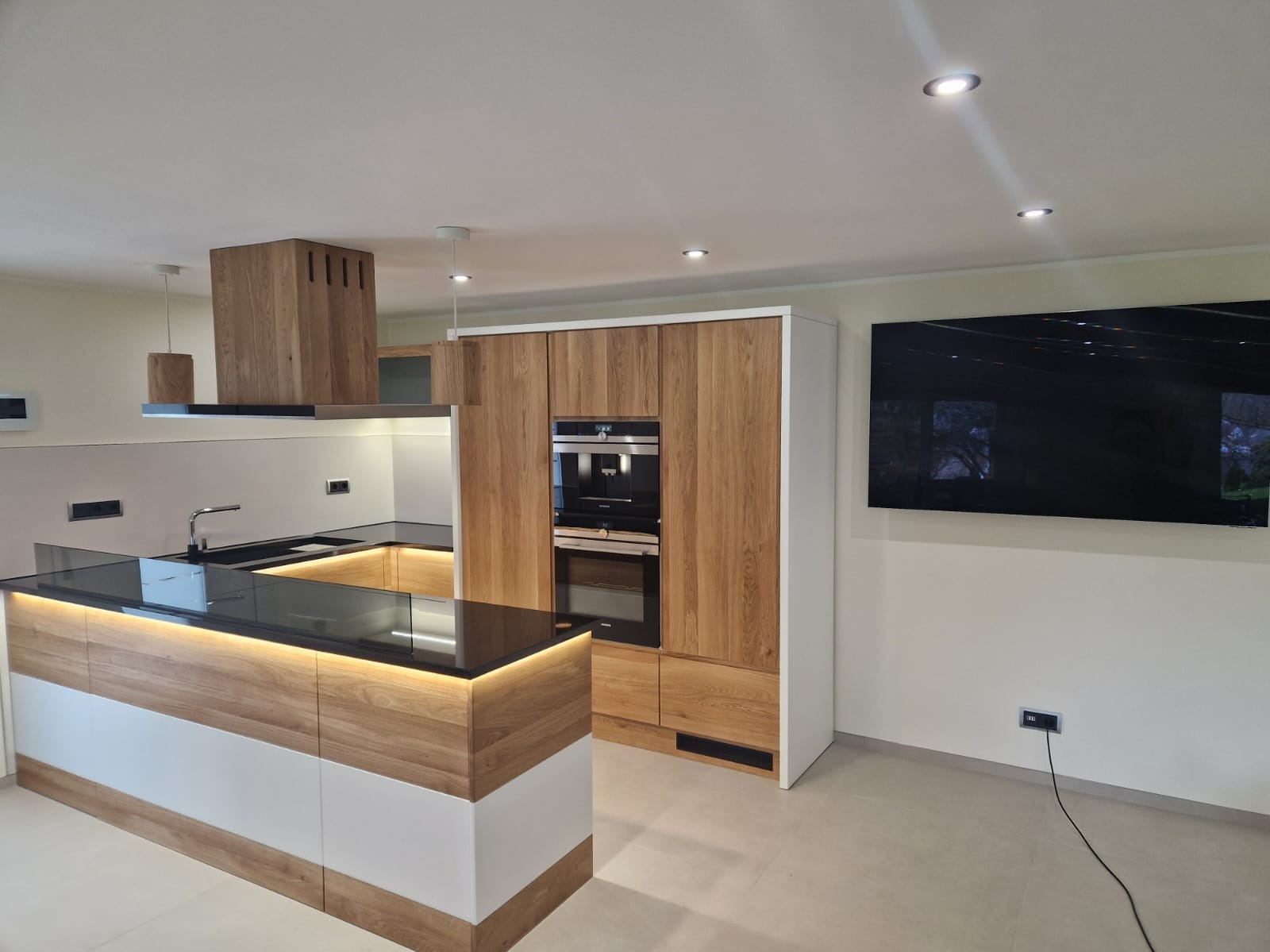 Nowoczesna kuchnia z wyspą, drewnianymi frontami, czarnym blatem i podświetleniem LED, zabudowana szafa z piekarnikiem i ekspresem do kawy, minimalistyczny okap.