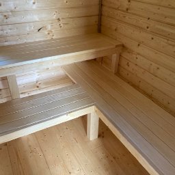 PW Pawelo Paweł Ostaszewski - Wnętrze sauny z jasnego drewna, ukazujące dwupoziomowe, narożne ławki i boazerię ścienną z widocznymi słojami drewna.
