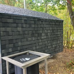 PW Pawelo Paweł Ostaszewski - Ściana budynku obłożona ciemnoszarą dachówką bitumiczną, widoczny fragment elewacji bocznej i rynna, w tle drzewo z czerwoną liną.