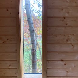 PW Pawelo Paweł Ostaszewski - Wnętrze drewnianego domu z wąskim oknem, przez które widać pień drzewa i jesienne liście.
