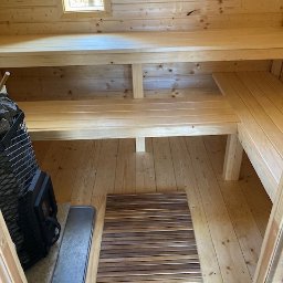 PW Pawelo Paweł Ostaszewski - Wnętrze sauny z drewnianymi ławkami, piecem z kamieniami i małym oknem, widok z perspektywy wejścia, na podłodze mata z drewnianych listewek.