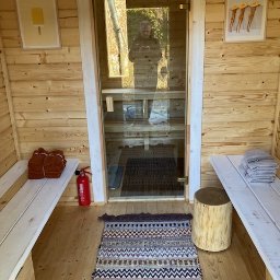 PW Pawelo Paweł Ostaszewski - Wnętrze drewnianej sauny z szklanymi drzwiami, widoczne drewniane ławki, dywanik w zygzaki, pień drzewa jako stolik oraz gaśnica. W odbiciu w szkle widoczna osoba robiąca zdjęcie.