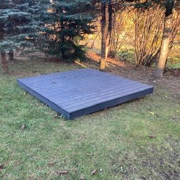 PW Pawelo Paweł Ostaszewski - Niska, szara platforma z desek kompozytowych na trawiastym terenie, otoczona drzewami w jesiennym słońcu.