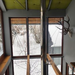 PW Pawelo Paweł Ostaszewski - Wnętrze domu z dużymi oknami, widok na zimowy krajobraz, dekoracja w postaci poroża na ścianie, metalowy komin na środku pomieszczenia.