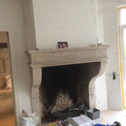 PW Pawelo Paweł Ostaszewski - Wnętrze w trakcie prac wykończeniowych: widoczny kamienny kominek, ściany przygotowane pod gładzie, narzędzia malarskie, wiadro z zaprawą i torba z materiałami budowlanymi. Na kominku stoi małe...