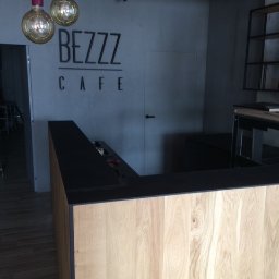 PW Pawelo Paweł Ostaszewski - Recepcja kawiarni BEZZZ CAFE z blatem w kolorze czarnym i frontem z jasnego drewna, oświetlona wiszącymi lampami z żarówkami Edisona.