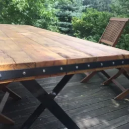 Stół ogrodowy z blatem z surowego drewna z metalowymi wzmocnieniami i nogami w kształcie litery X, na drewnianym tarasie, z krzesłami w tle.