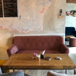 PW Pawelo Paweł Ostaszewski - Wnętrze kawiarni: rustykalny stół z drewna i metalu, różowa sofa z guzikami, szary fotel, menu na tablicy i dekoracyjna ściana z efektem postarzania. Na stole deser z owocami, piwo i popielniczka.