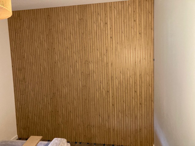 Ściana wykończona pionowymi panelami drewnopodobnymi, widoczne fragmenty podłogi z panelami i materiałami budowlanymi.