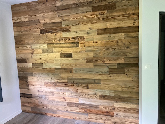 Ściana wykończona panelami z postarzanego drewna o zróżnicowanej kolorystyce, widoczne sęki i usłojenie, jasne tło podłogi i białe ściany boczne.
