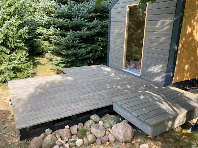 Szara drewniana podłoga tarasu przed domkiem z szarą elewacją, widok z niskiej perspektywy na tle zieleni drzew, kamienie dekoracyjne u podstawy tarasu.