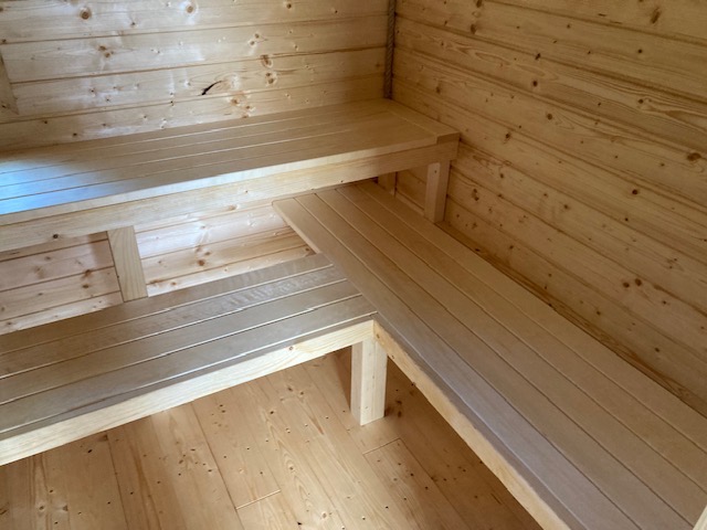 Wnętrze sauny z jasnego drewna, ukazujące dwupoziomowe, narożne ławki i boazerię ścienną z widocznymi słojami drewna.