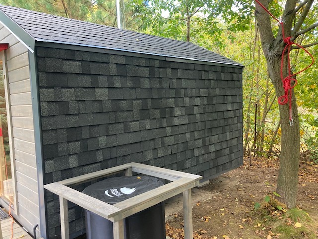 Ściana budynku obłożona ciemnoszarą dachówką bitumiczną, widoczny fragment elewacji bocznej i rynna, w tle drzewo z czerwoną liną.