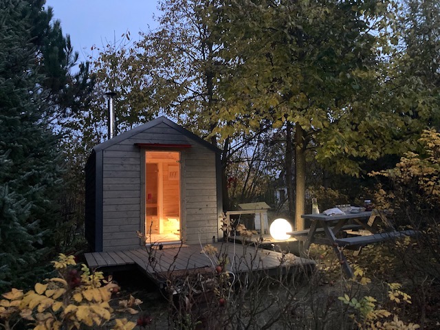 Mały, drewniany domek z kominem, prawdopodobnie sauna, z otwartymi drzwiami, oświetlony od wewnątrz, na drewnianym podeście, obok stolik piknikowy i oświetlona kula, otoczony drzewami i krzewami...