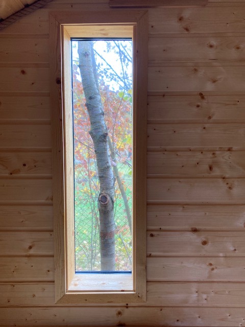 Wnętrze drewnianego domu z wąskim oknem, przez które widać pień drzewa i jesienne liście.