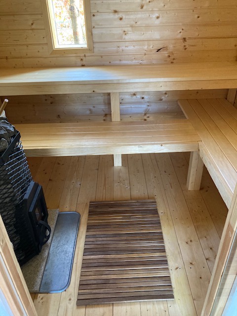 Wnętrze sauny z drewnianymi ławkami, piecem z kamieniami i małym oknem, widok z perspektywy wejścia, na podłodze mata z drewnianych listewek.