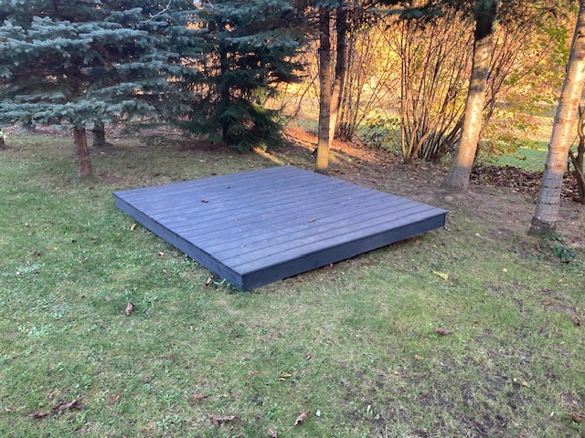 Niska, szara platforma z desek kompozytowych na trawiastym terenie, otoczona drzewami w jesiennym słońcu.
