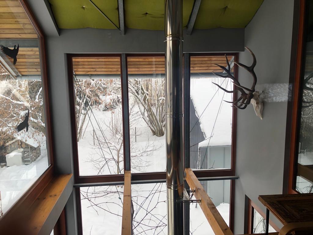 Wnętrze domu z dużymi oknami, widok na zimowy krajobraz, dekoracja w postaci poroża na ścianie, metalowy komin na środku pomieszczenia.