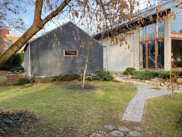 Nowoczesny dom z ciemnoszarą dobudówką, widok z zewnątrz, ścieżka z kamieni prowadzi do przeszklonej części domu, drzewo z opadłymi liśćmi w pierwszym planie.