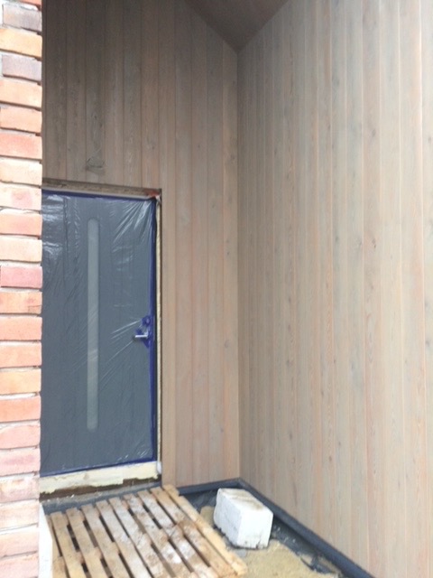 Wejście do budynku w trakcie wykańczania: drzwi zabezpieczone folią, ściany wyłożone jasnym drewnem, paleta na podłodze, widoczny fragment muru z cegły.