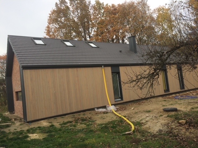 Nowoczesny dom jednorodzinny z drewnianą elewacją i grafitowym dachem z oknami połaciowymi, widoczny żółty wąż doprowadzający wodę.